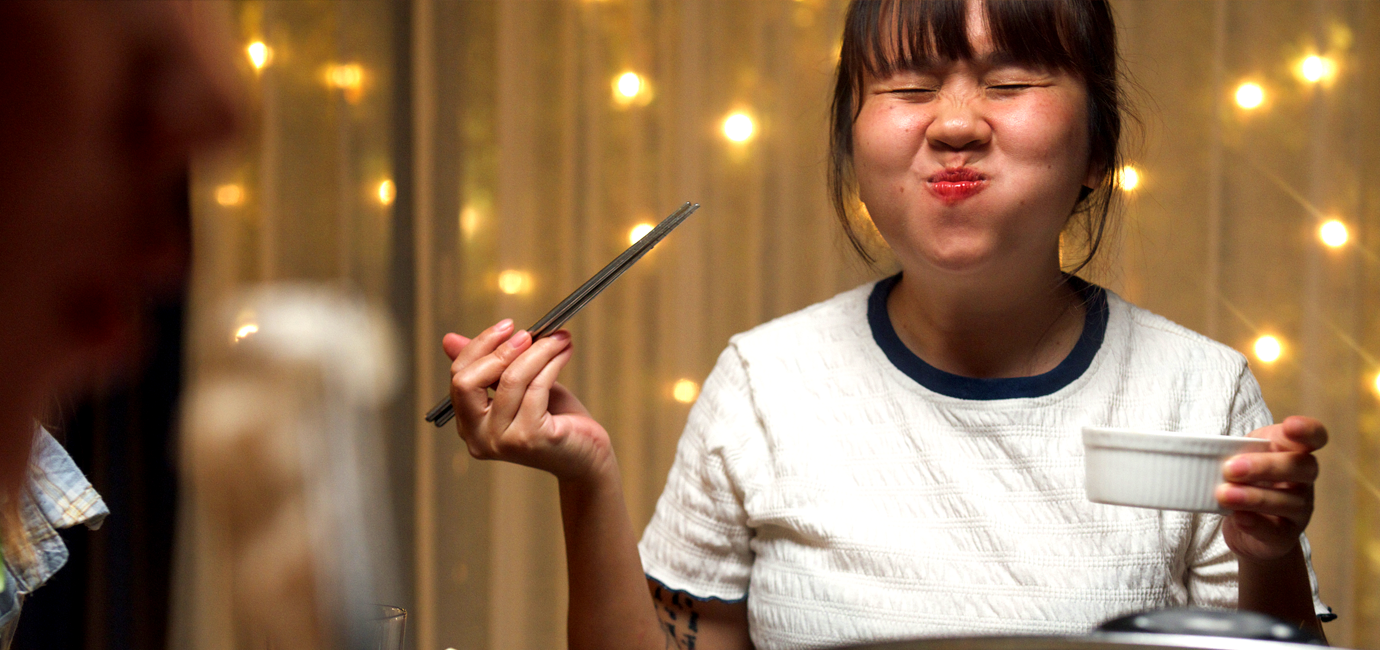 A happy woman holding chopsticks and a bowl, delighting in the food she is eating, with a curtain and twinkling, yellow lights in the background.