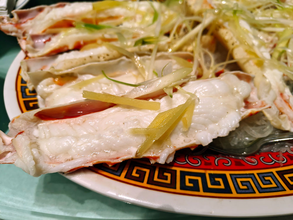 An image of steamed king crab with vegetables atop glass noodles, on a golden, black, red, and white dish.
